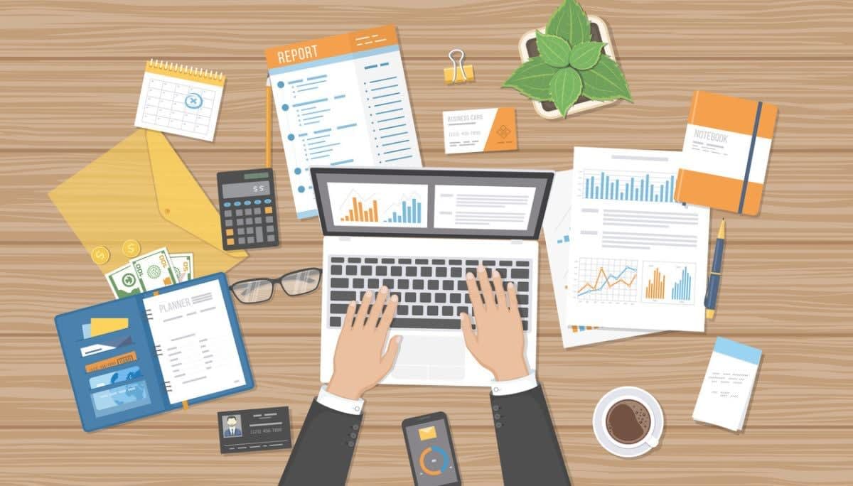 Overhead view of hands typing on a laptop surrounded by reports, charts, a planner, and office tools, symbolizing strategic planning and business analysis.